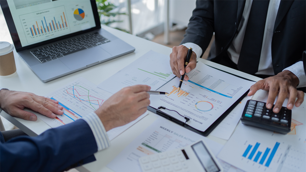 Two people in suits analyzing financial reports with a laptop and calculator at a desk.