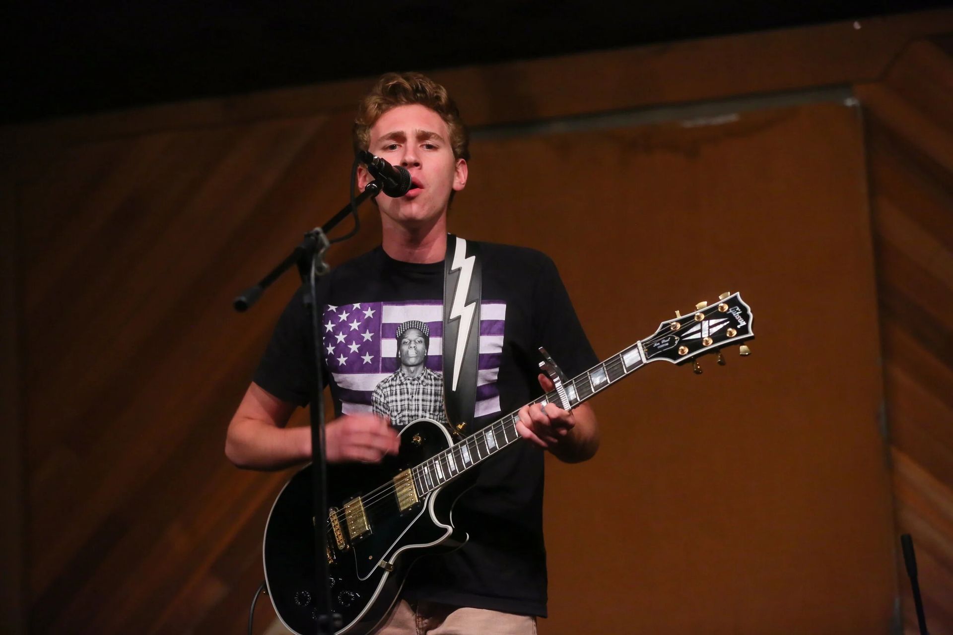 Man playing a black guitar and singing into a microphone on stage, wearing a t-shirt with a purple and white design.