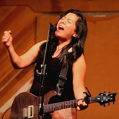 Woman singing and playing electric guitar on stage, arm raised, mouth open.