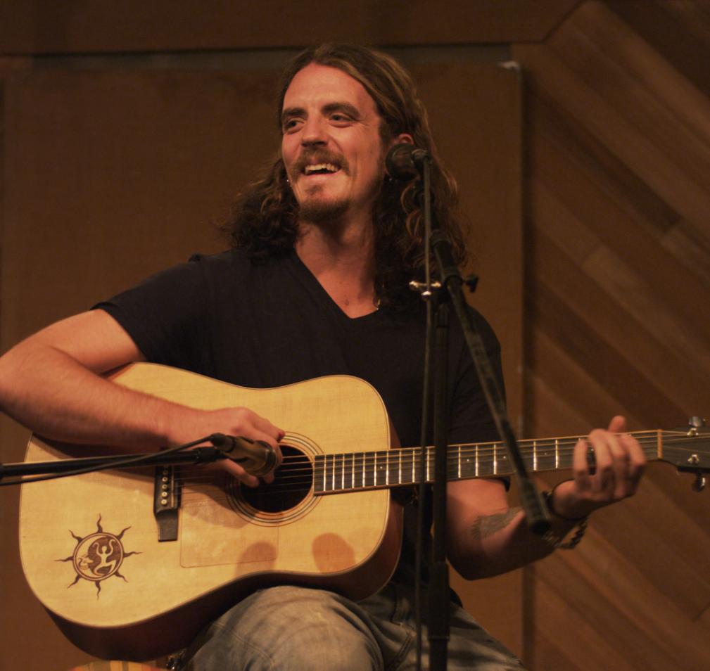 Man with long hair playing acoustic guitar and singing into a microphone. Smiling, seated. Rustic interior.