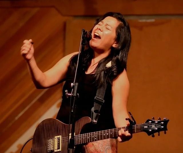 Woman singing and playing electric guitar on stage, arm raised, mouth open, black top.