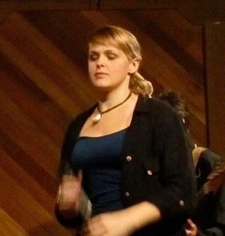 Woman wearing a blue top and black jacket, standing in front of a wood-paneled wall.