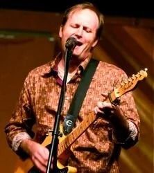 Man singing, playing electric guitar on stage, eyes closed, wearing patterned brown shirt.