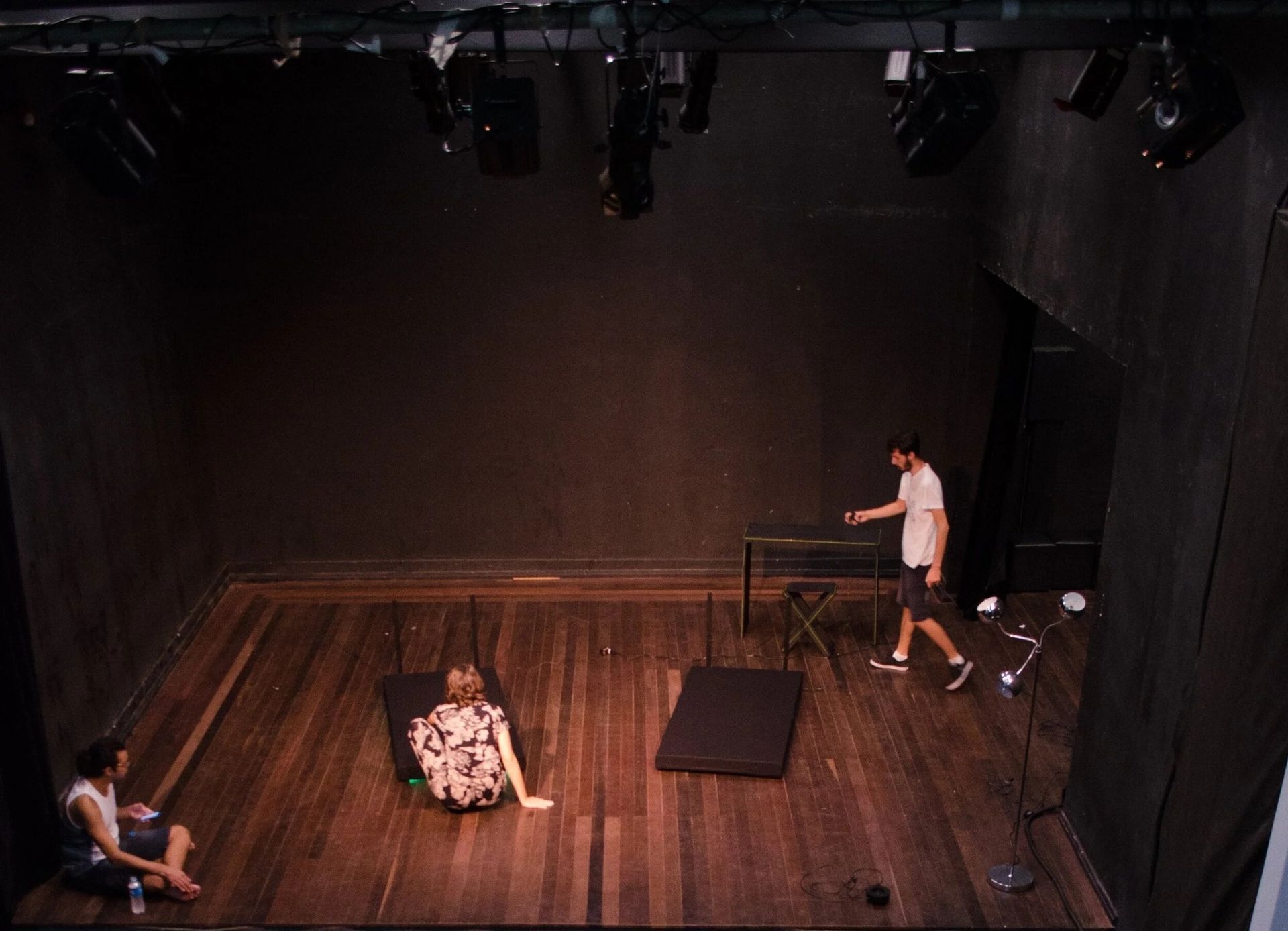 A dimly lit theater stage; two people seated, one lying down. A third person walks, pointing.