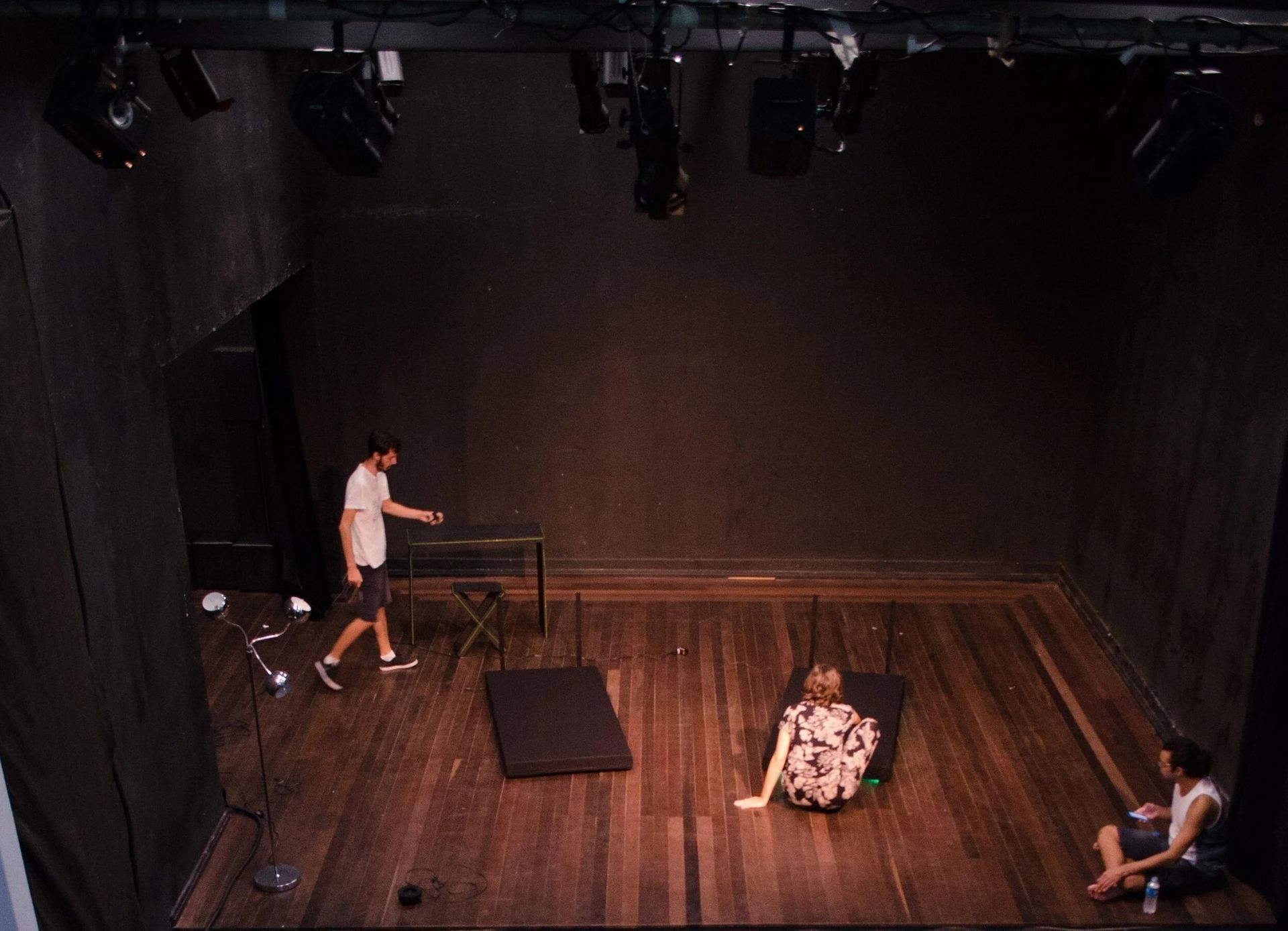 A theater stage with three people. One stands, another lies down, and the third sits. Black backdrop.