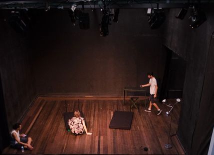 A dimly lit theater stage; two people seated, one lying down. A third person walks, pointing.