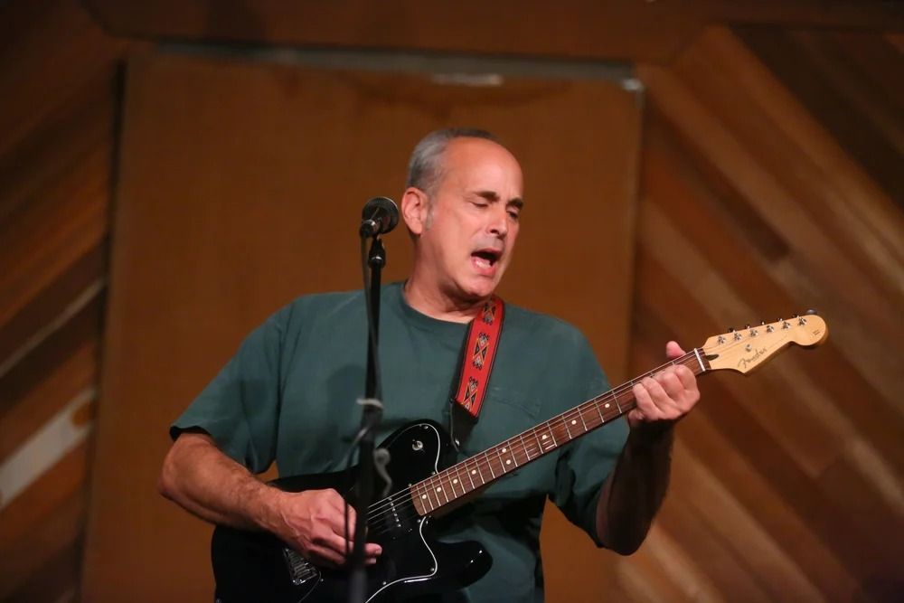 Man playing black electric guitar and singing into a microphone on stage, wearing a green shirt.