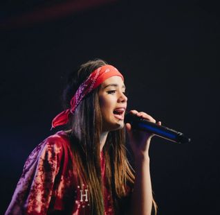 Woman singing into a microphone, wearing a red headband and shirt on a dark stage.