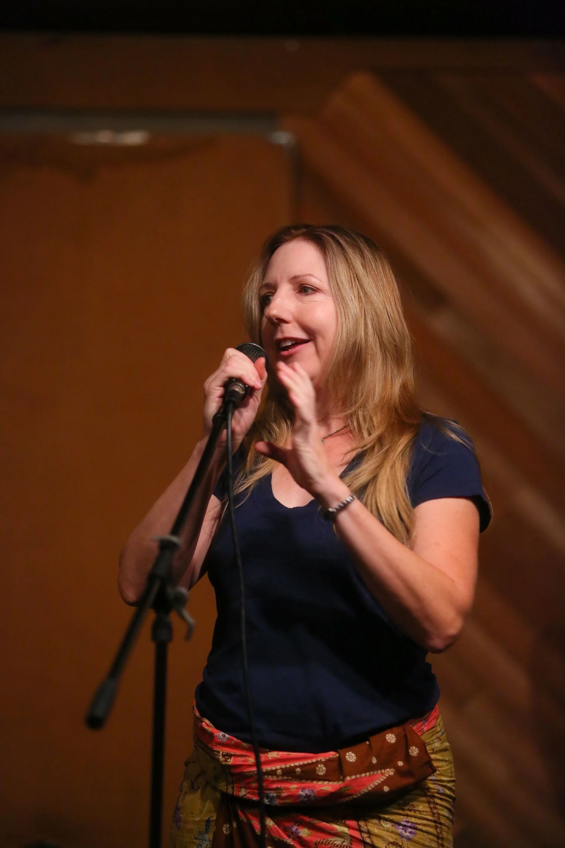 Woman with long blonde hair, singing into a microphone. She wears a blue shirt and patterned skirt. Inside, warm lighting.