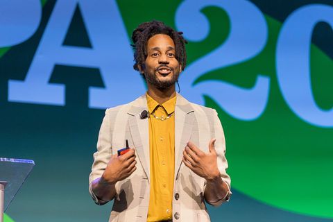 A man wearing a yellow shirt and a white jacket is giving a speech.