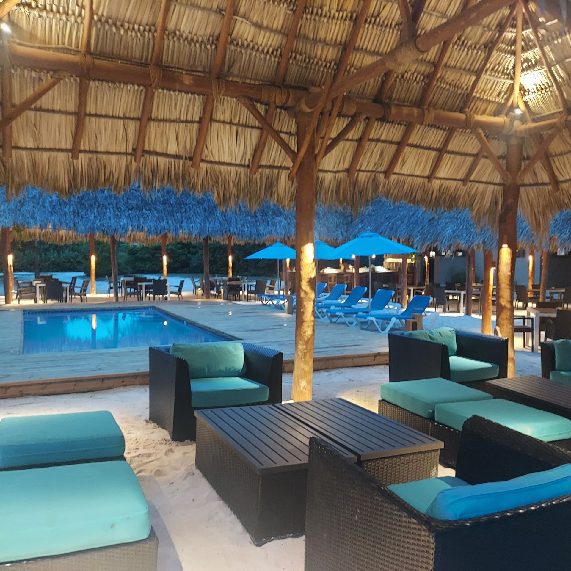Lounge area with a pool and thatched roof structures; teal cushions, blue umbrellas.