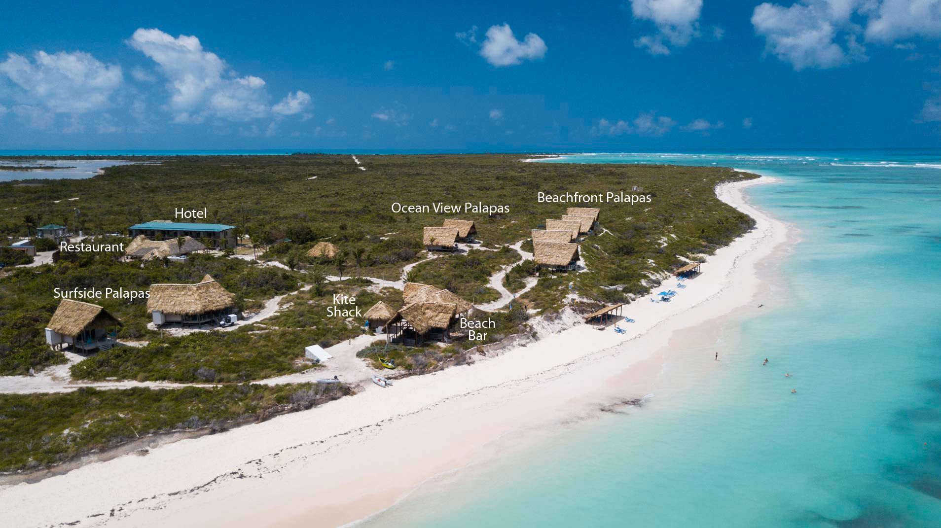Aerial view of a tropical island with thatch-roofed cabins along a white sand beach and turquoise water.