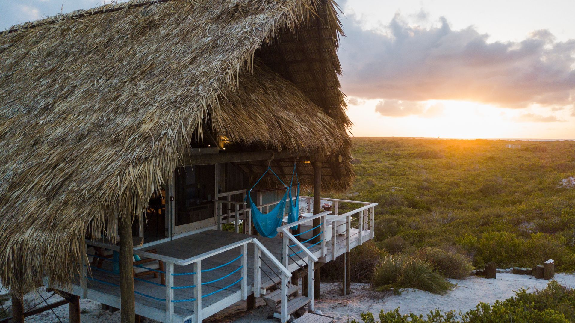 Thatched-roof cabin on a white-sand beach at sunset; blue hammock hangs on the porch.