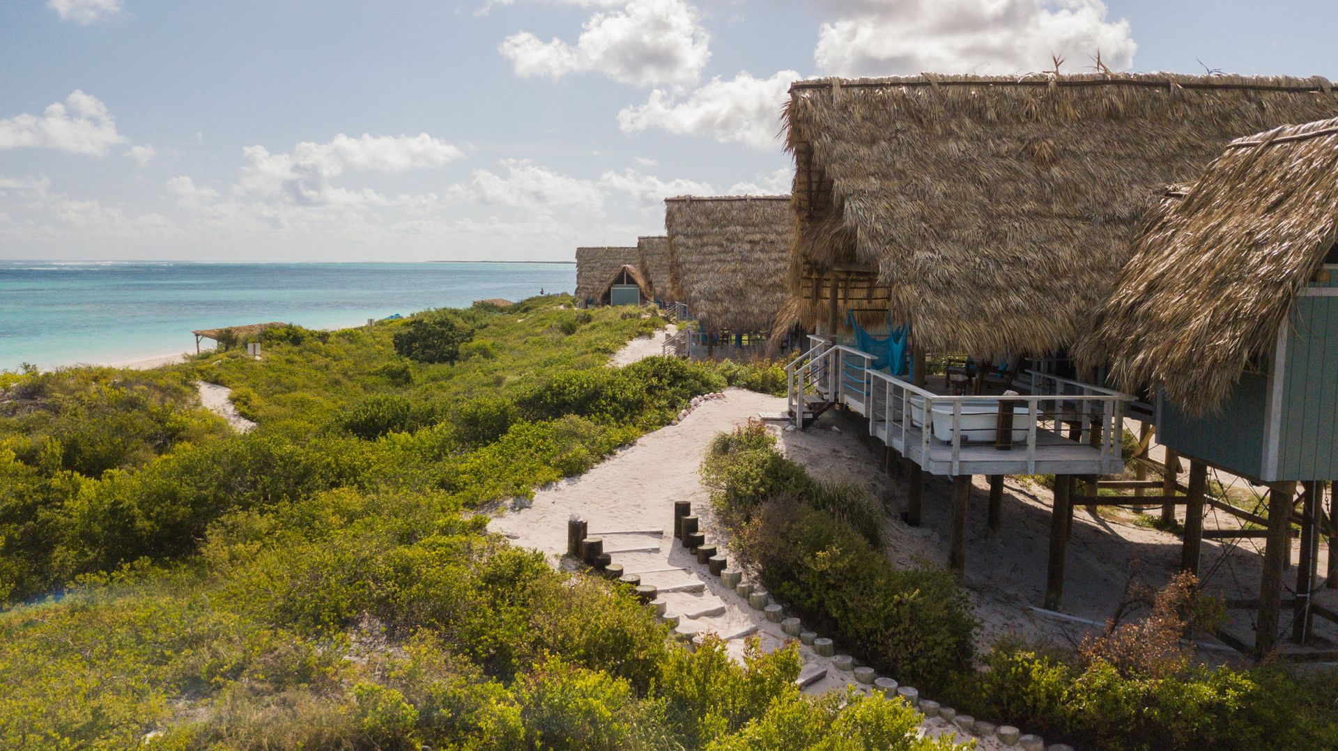 Beachfront huts with thatched roofs on stilts; sand path, ocean view, sunny day.