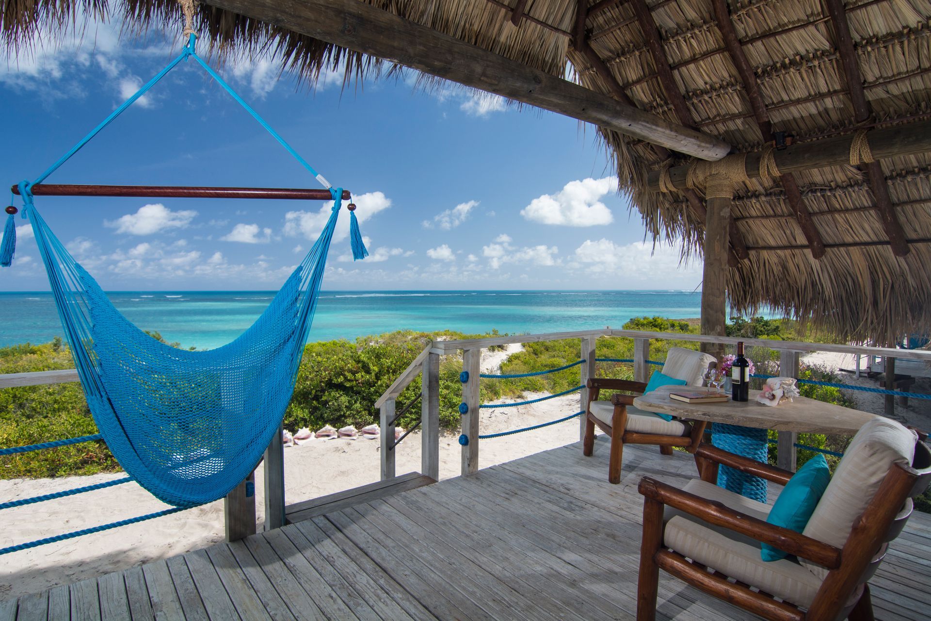 Blue hammock on a wooden deck overlooking a turquoise ocean, with table and chairs.