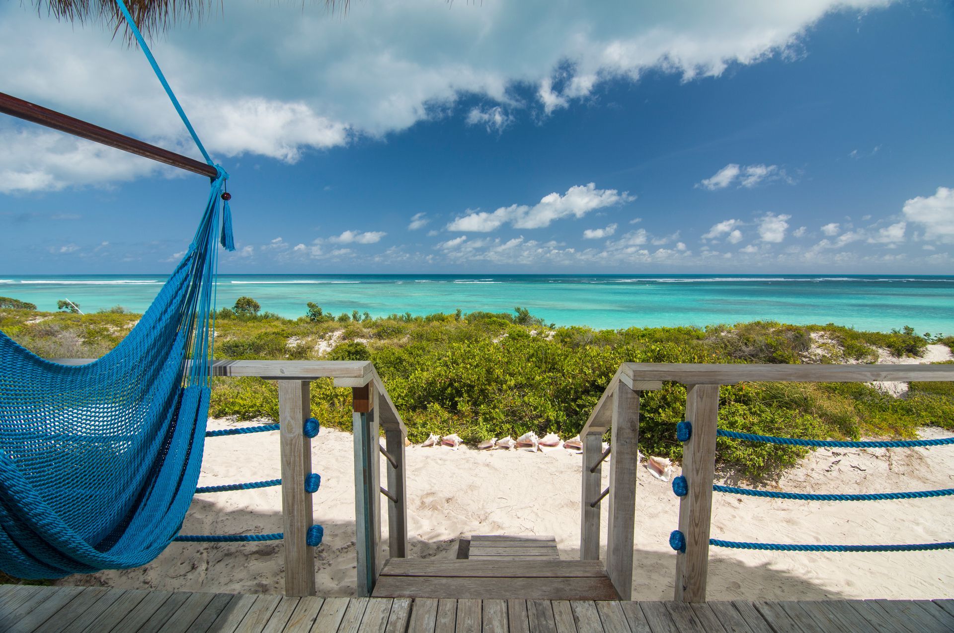 Blue hammock on a deck overlooking a sandy beach, turquoise ocean, and blue sky with clouds.
