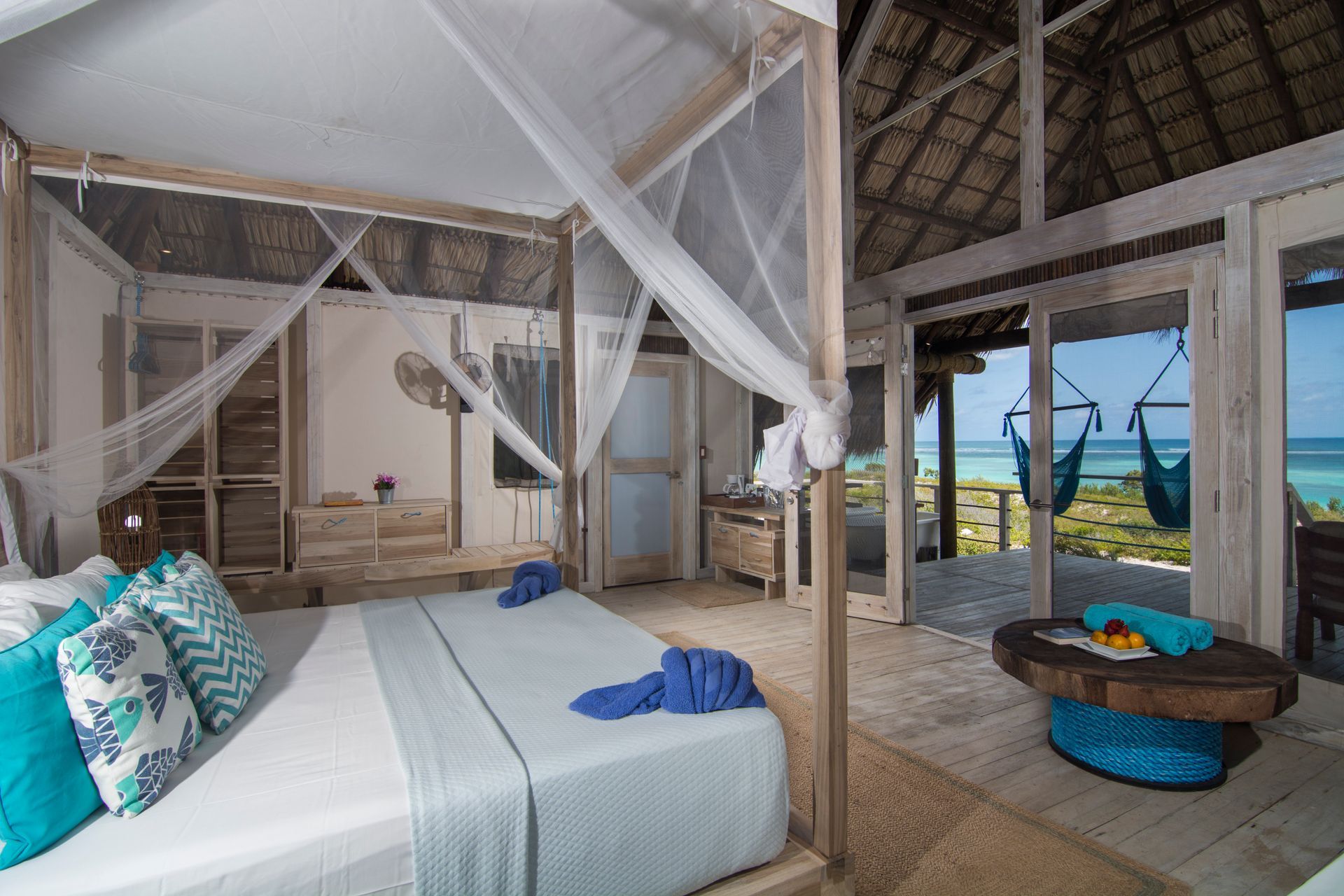 Bedroom with four-poster bed, mosquito net, open doors to beach, turquoise accents, wooden decor.