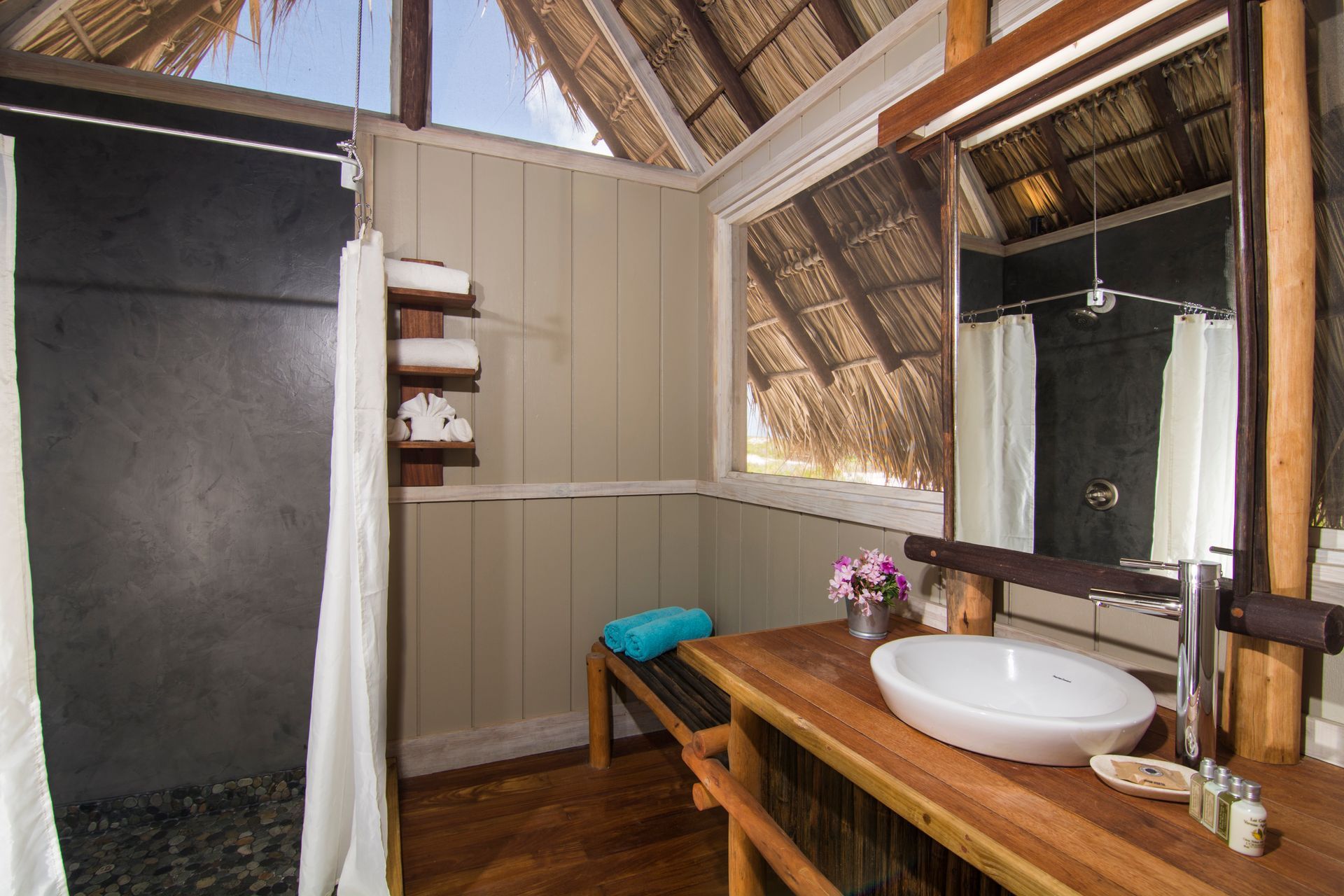 Bathroom with wooden vanity, white sink, shower, and open-air thatched roof.