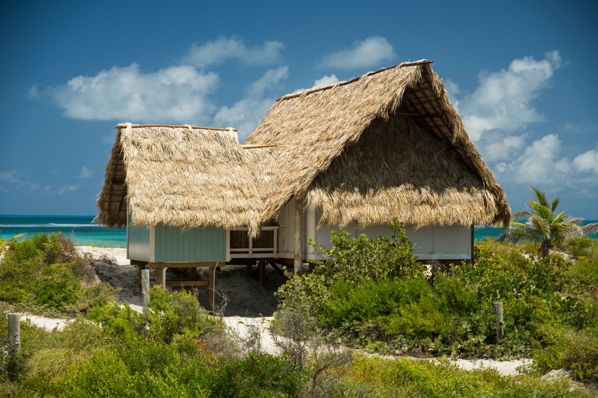 Thatched-roof cottage on stilts; beach setting with turquoise water and blue sky.