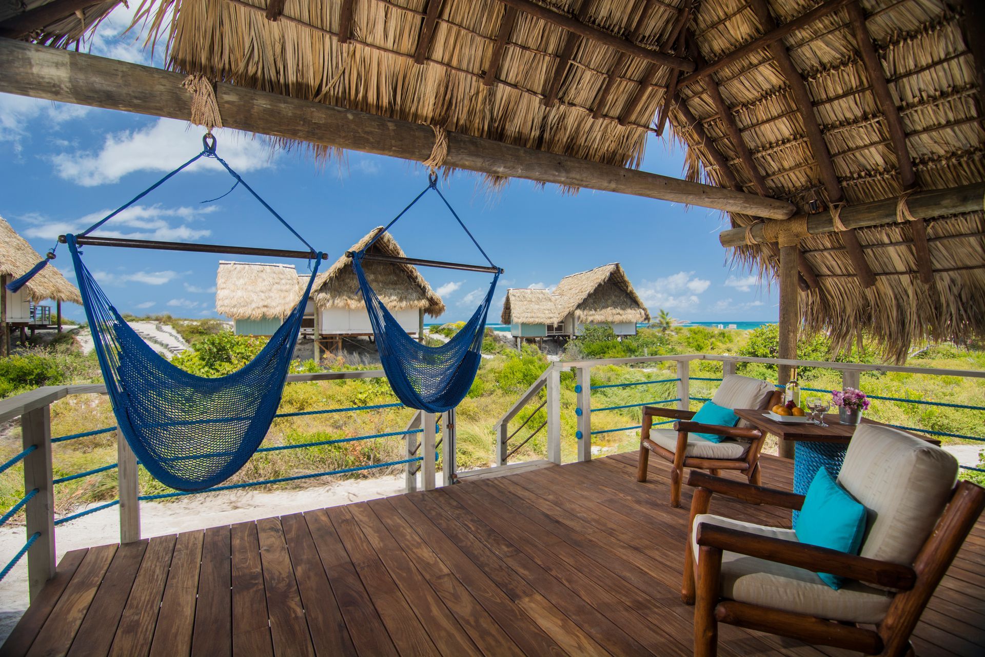 Wooden deck with two blue hammocks, chairs, and a view of thatched-roof bungalows near the sea under a blue sky.