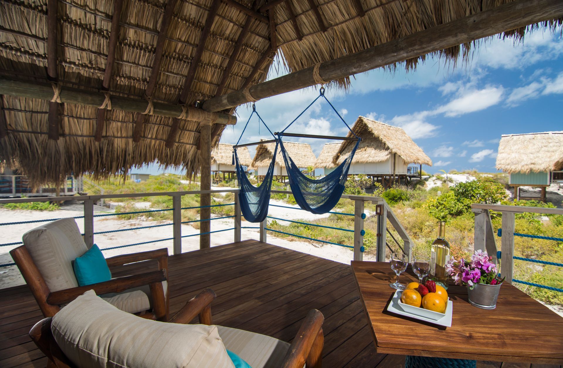 Wooden patio with chairs, hammock, and table set with fruit and flowers overlooking beach huts.
