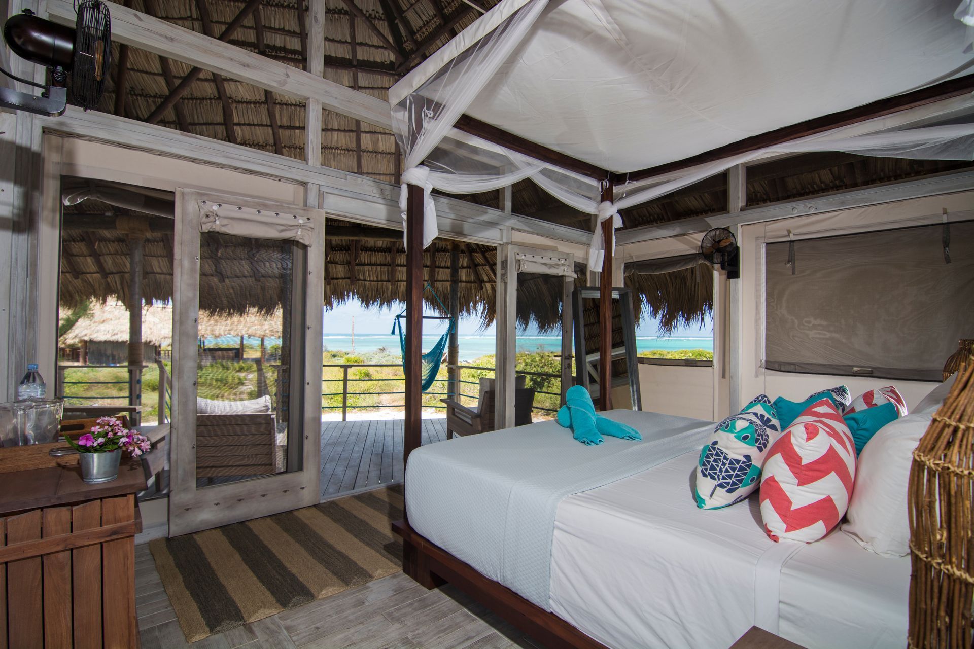 Bedroom with thatched roof, open doors to a balcony overlooking the ocean. Bed with pillows and mosquito net.