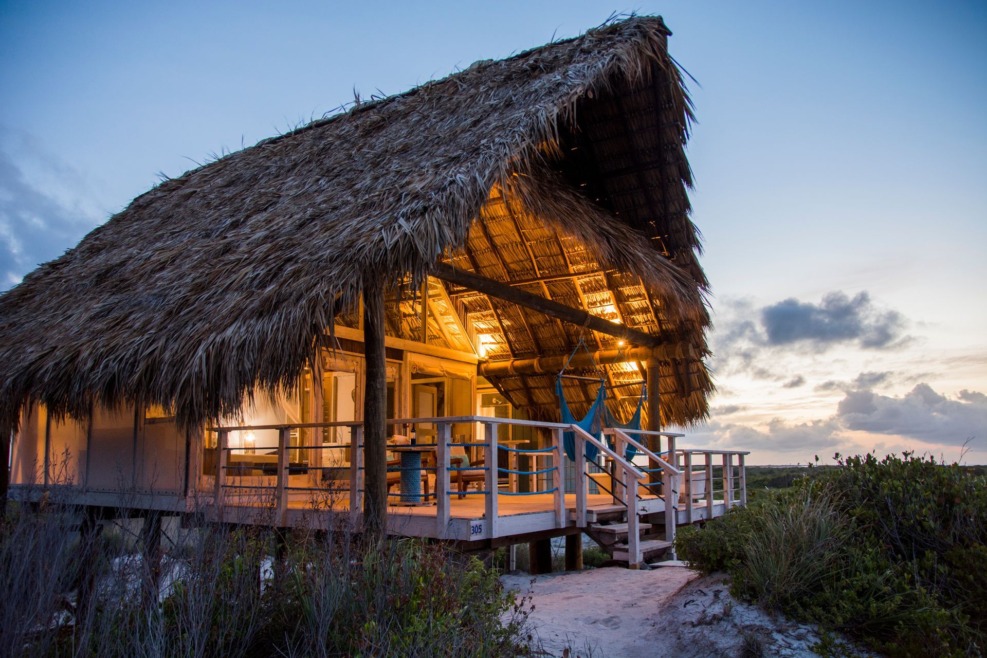 Rustic cabin with thatched roof lit from within at dusk, on a raised wooden deck.