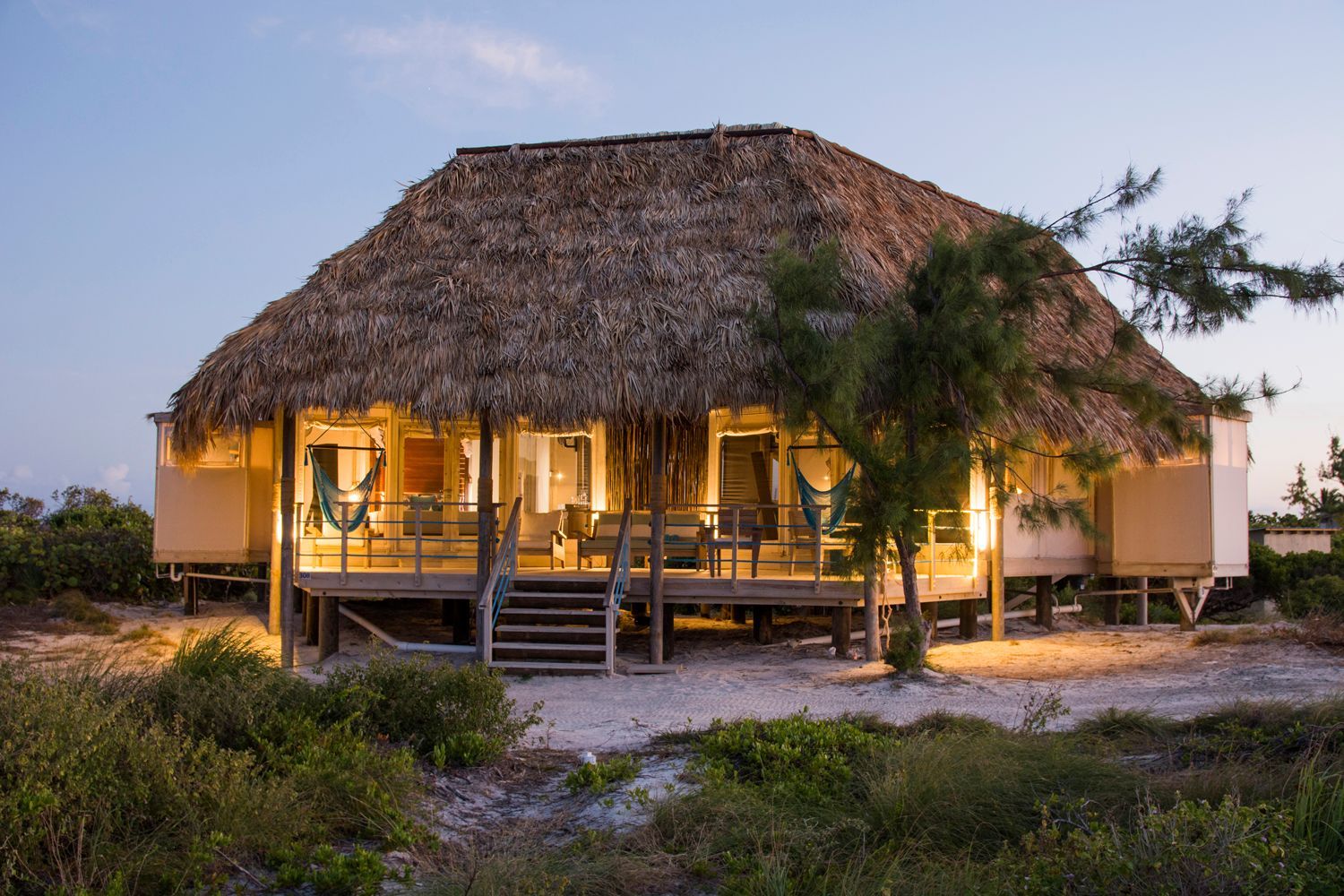 Cottage with thatched roof and illuminated interior, set on wooden stilts, surrounded by vegetation at dusk.