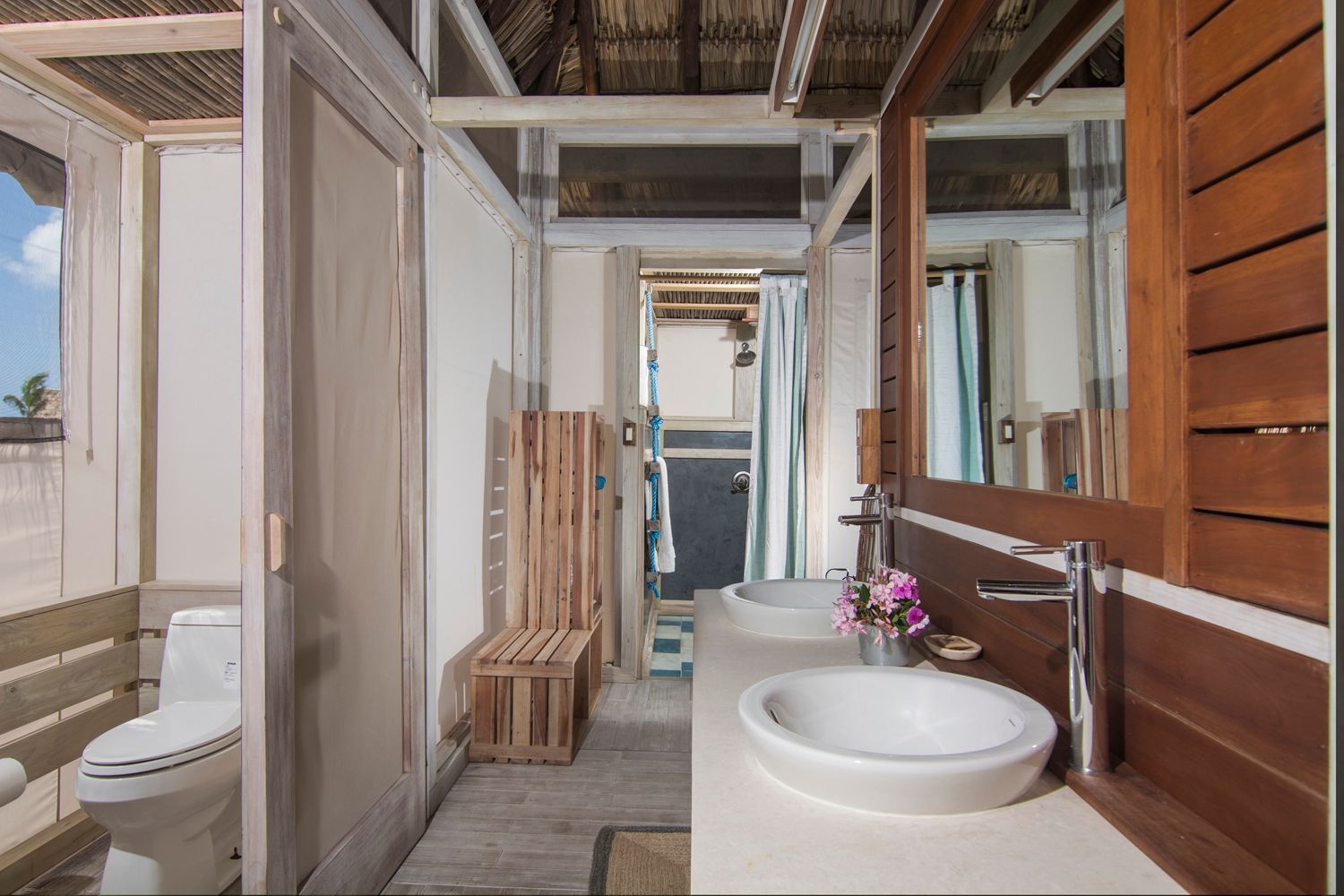 Rustic bathroom with a toilet, double sinks, and an open-air shower, mostly white and wood-toned.