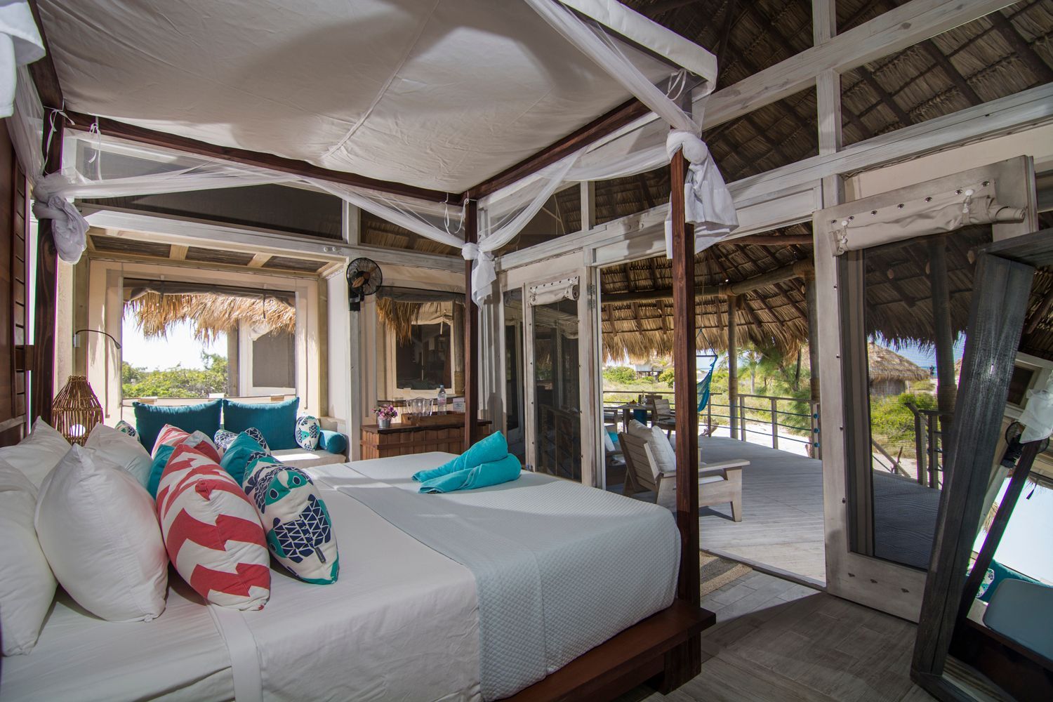 Bedroom with canopy bed, turquoise and coral pillows, thatched roof, and open to a deck.