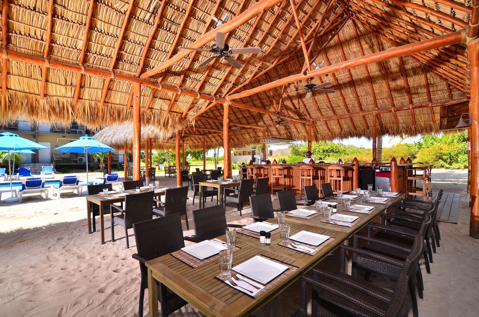 Outdoor dining area with tables set for a meal under a thatched roof on a sandy beach.