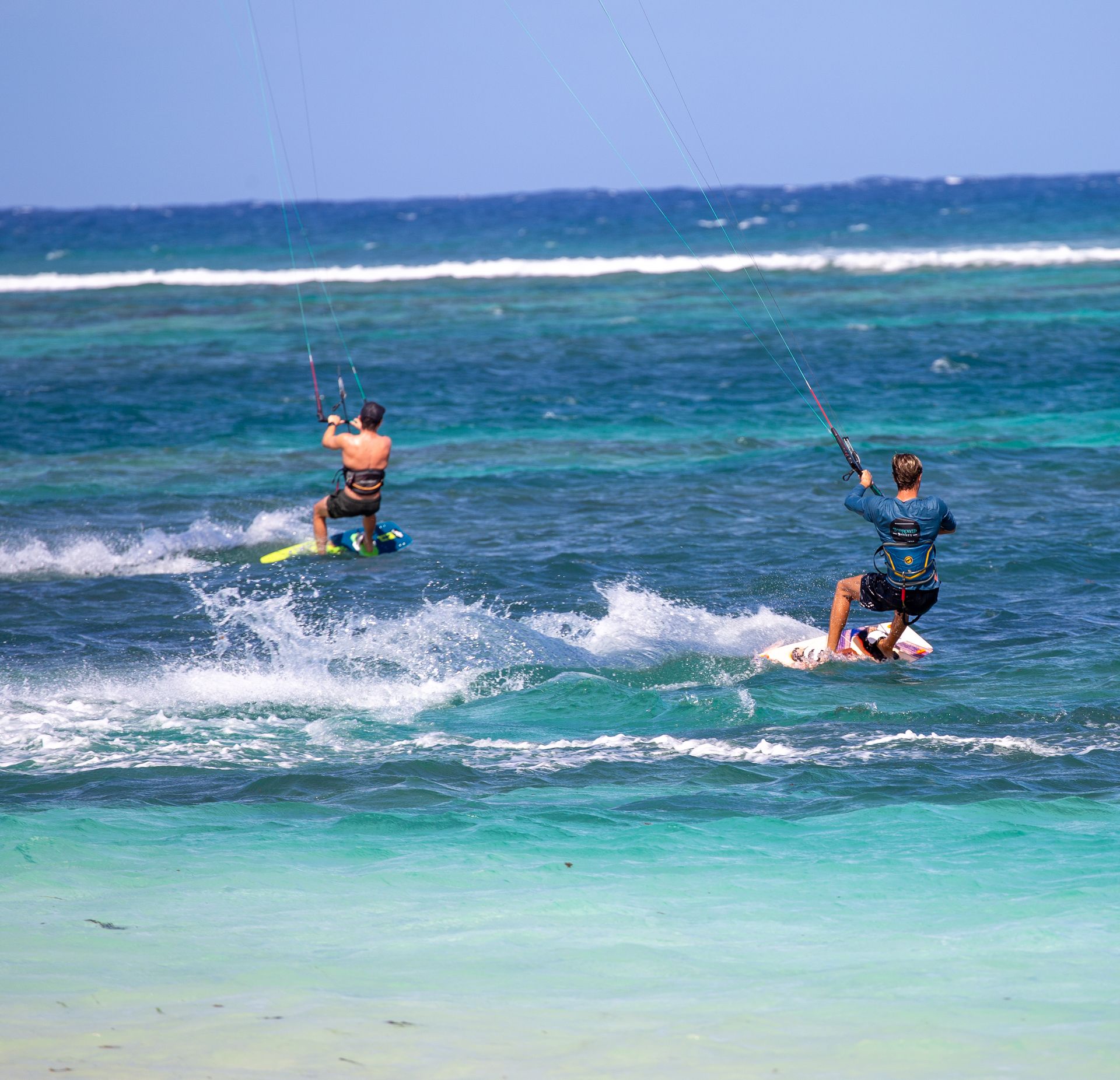 Two kite surfers on turquoise water near a beach. Blue sky overhead.