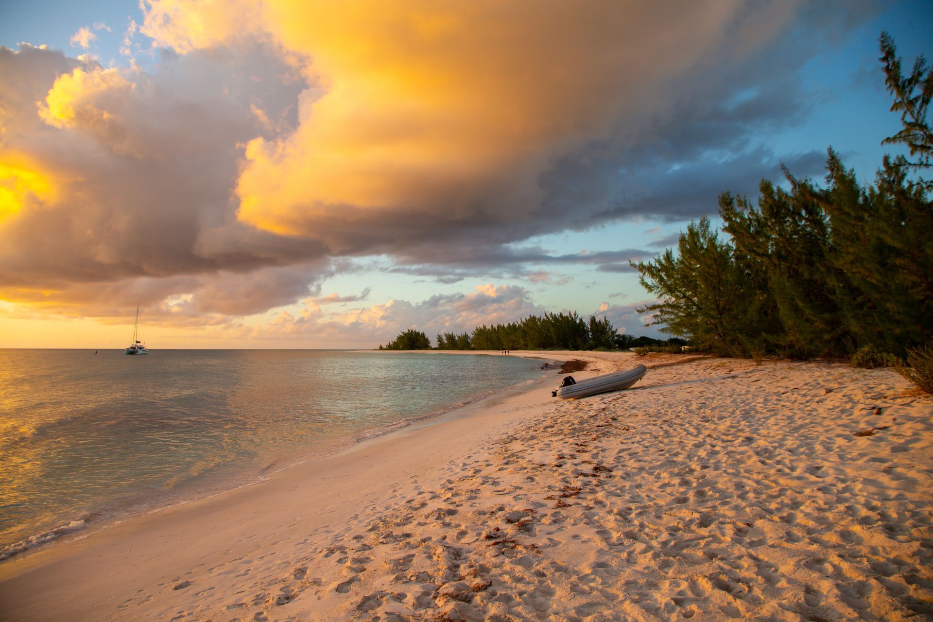 Beach at sunset: golden clouds, calm water, white sand, trees on the right, small boat in the distance.