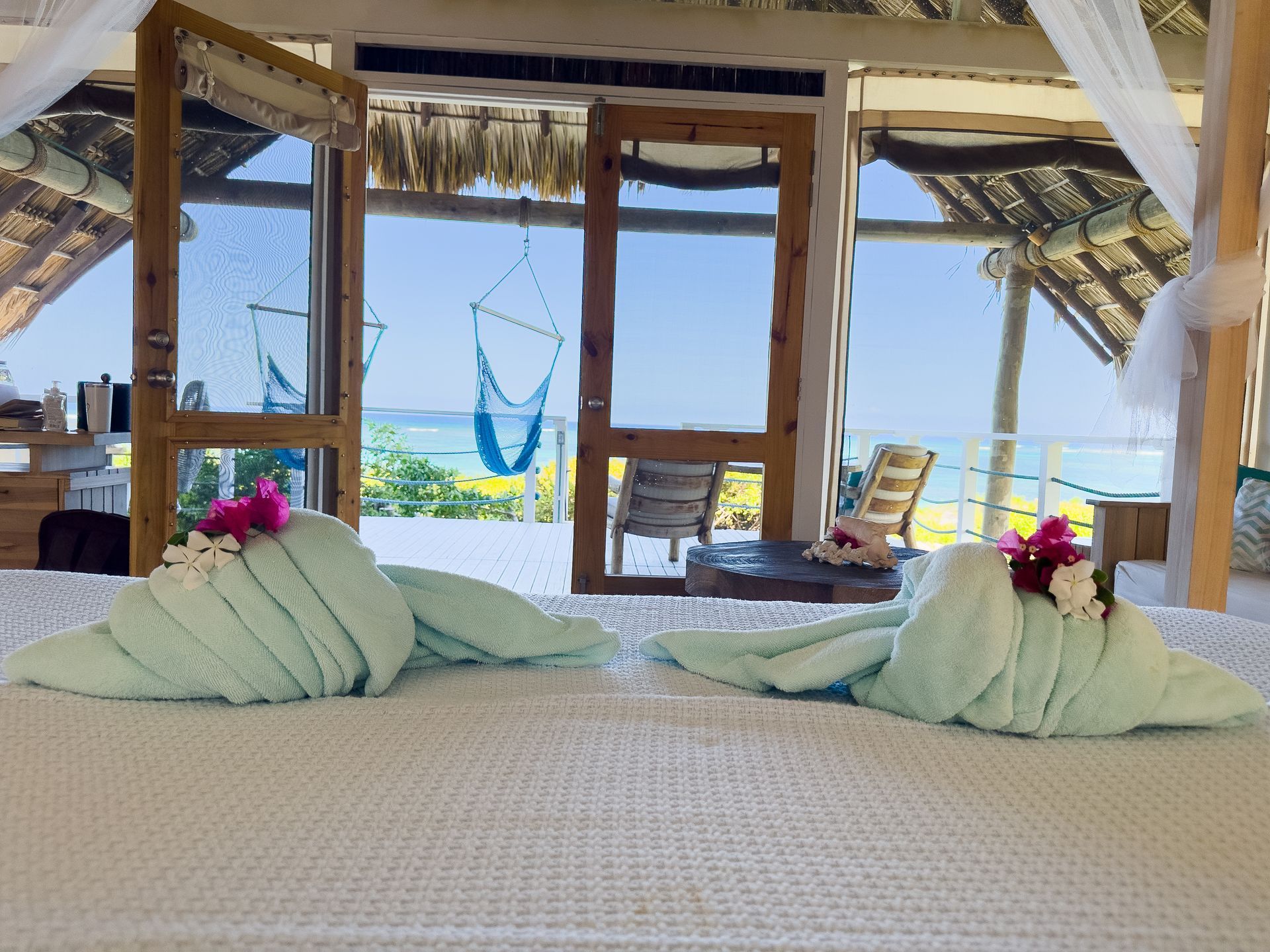 Bed decorated with towel swans and flowers, overlooking ocean through open doors.