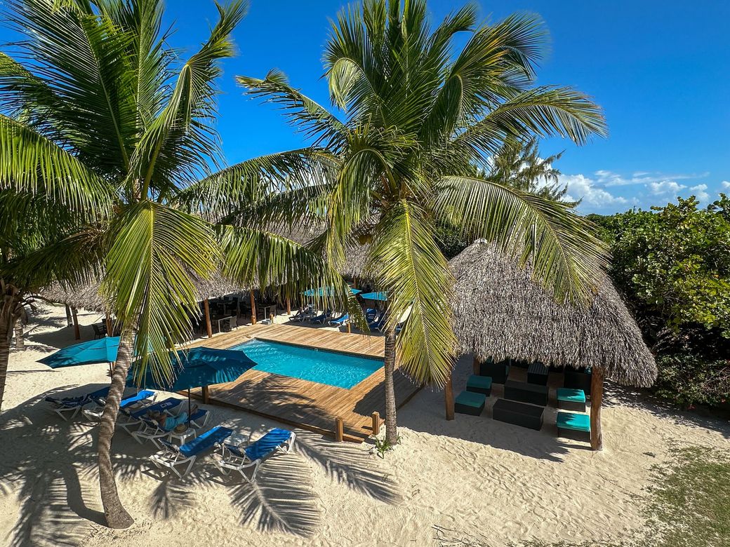 Tropical beach scene with pool, thatched roofs, palm trees, and lounge chairs on sand.