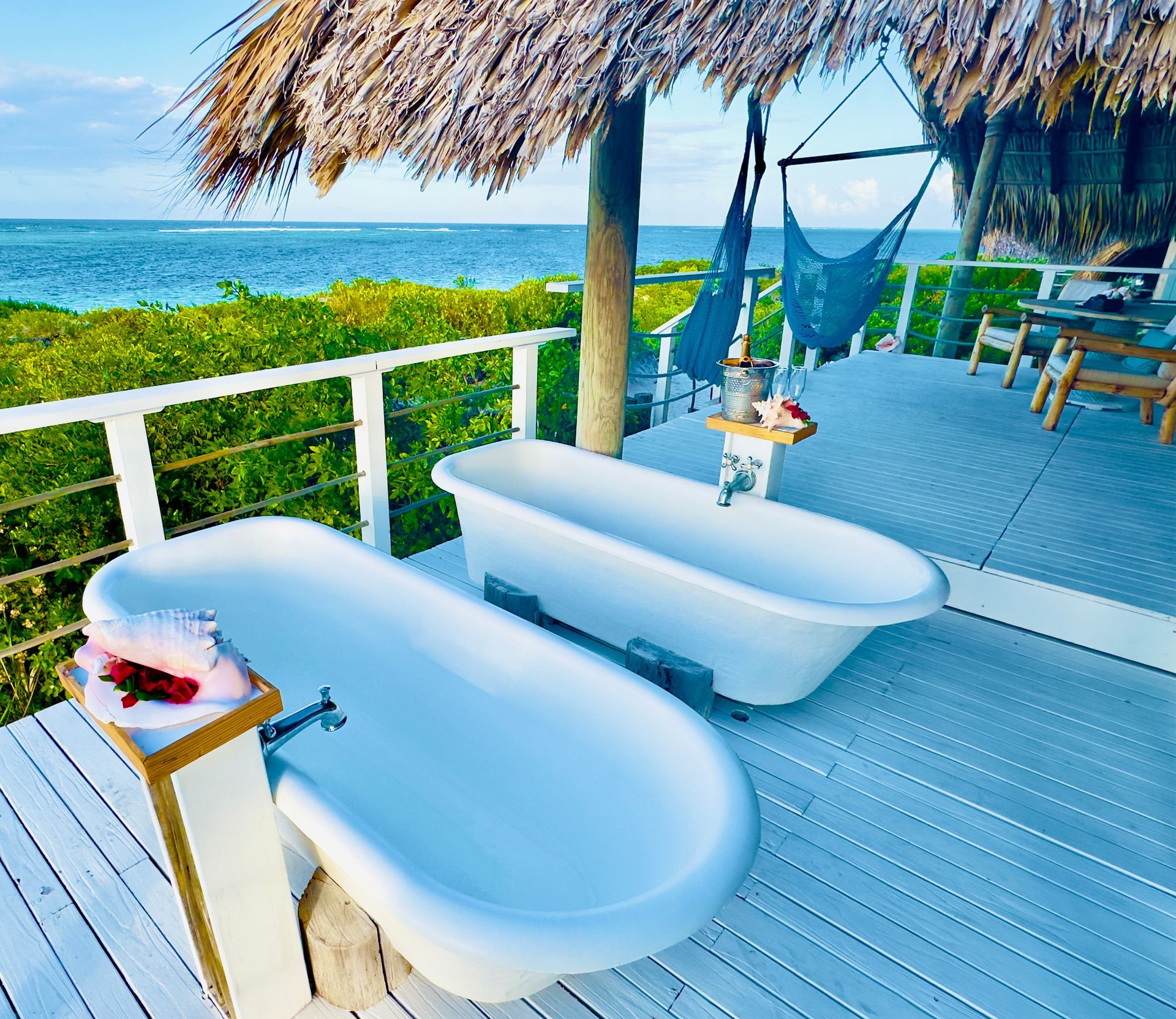 Two white bathtubs on a wooden deck overlooking the ocean under a thatched roof.