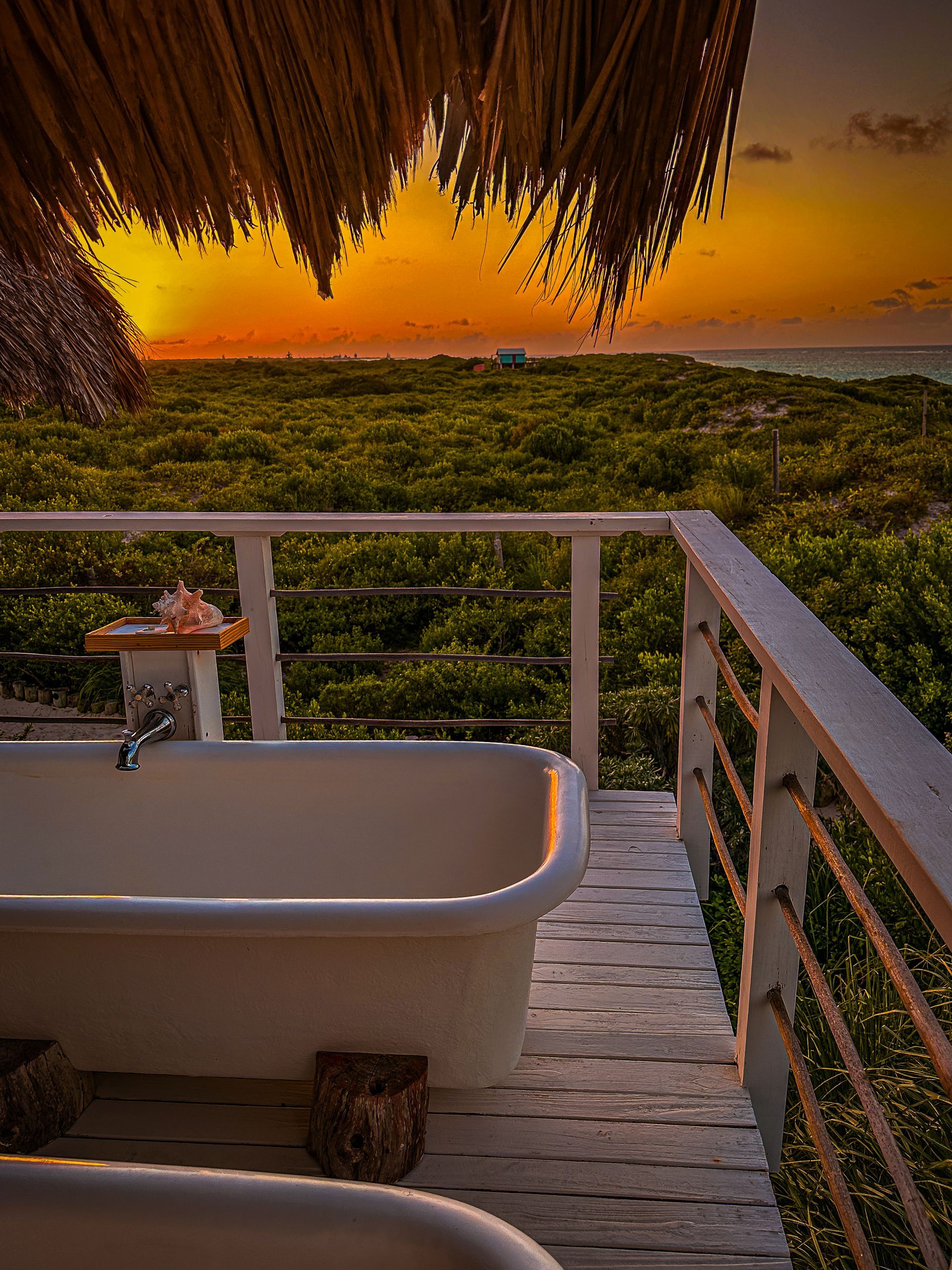 Sunset view from a wooden balcony with a bathtub overlooking lush greenery and the sea.