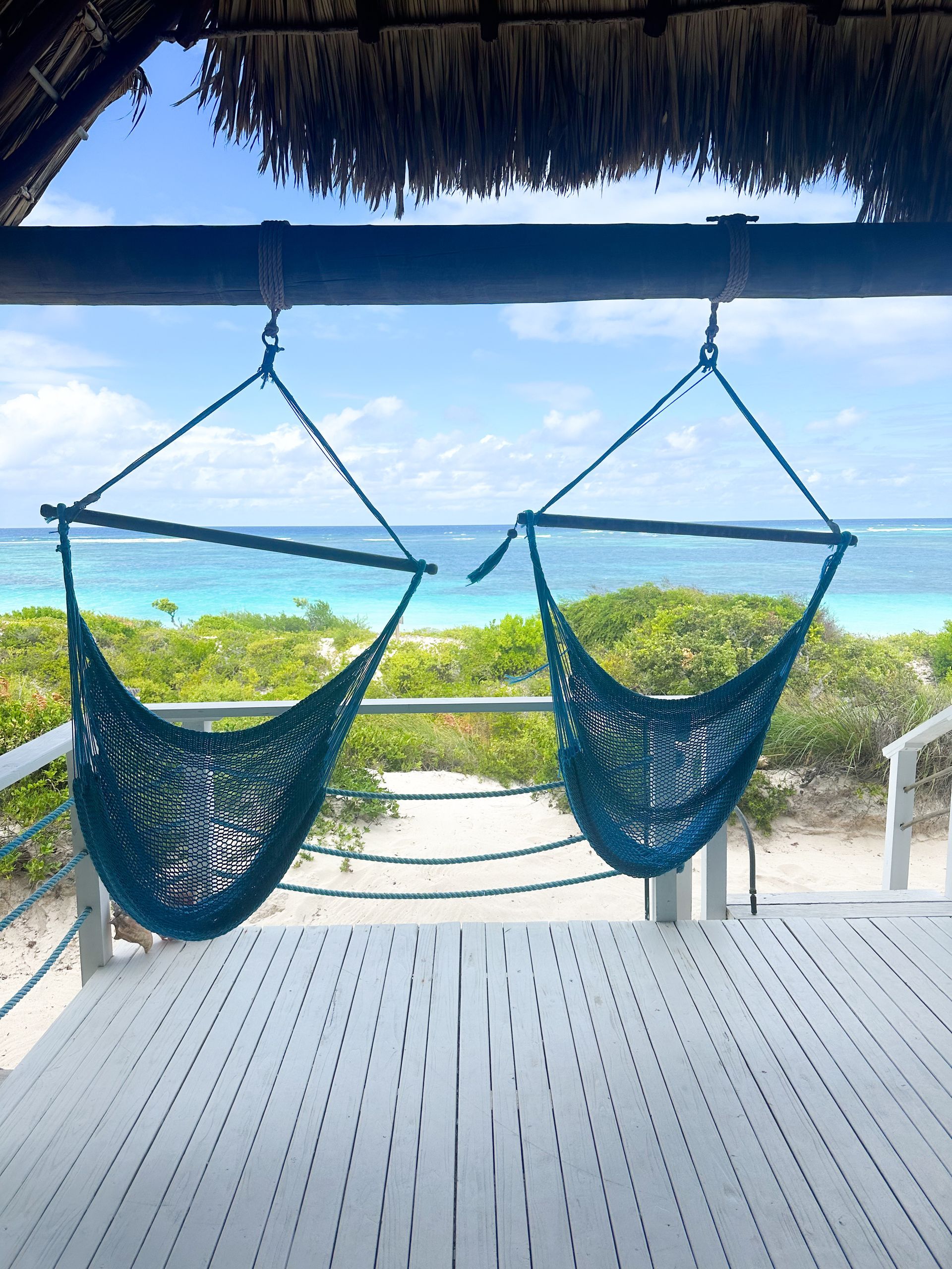 Two teal hammock chairs hang on a deck, overlooking a turquoise ocean and blue sky.
