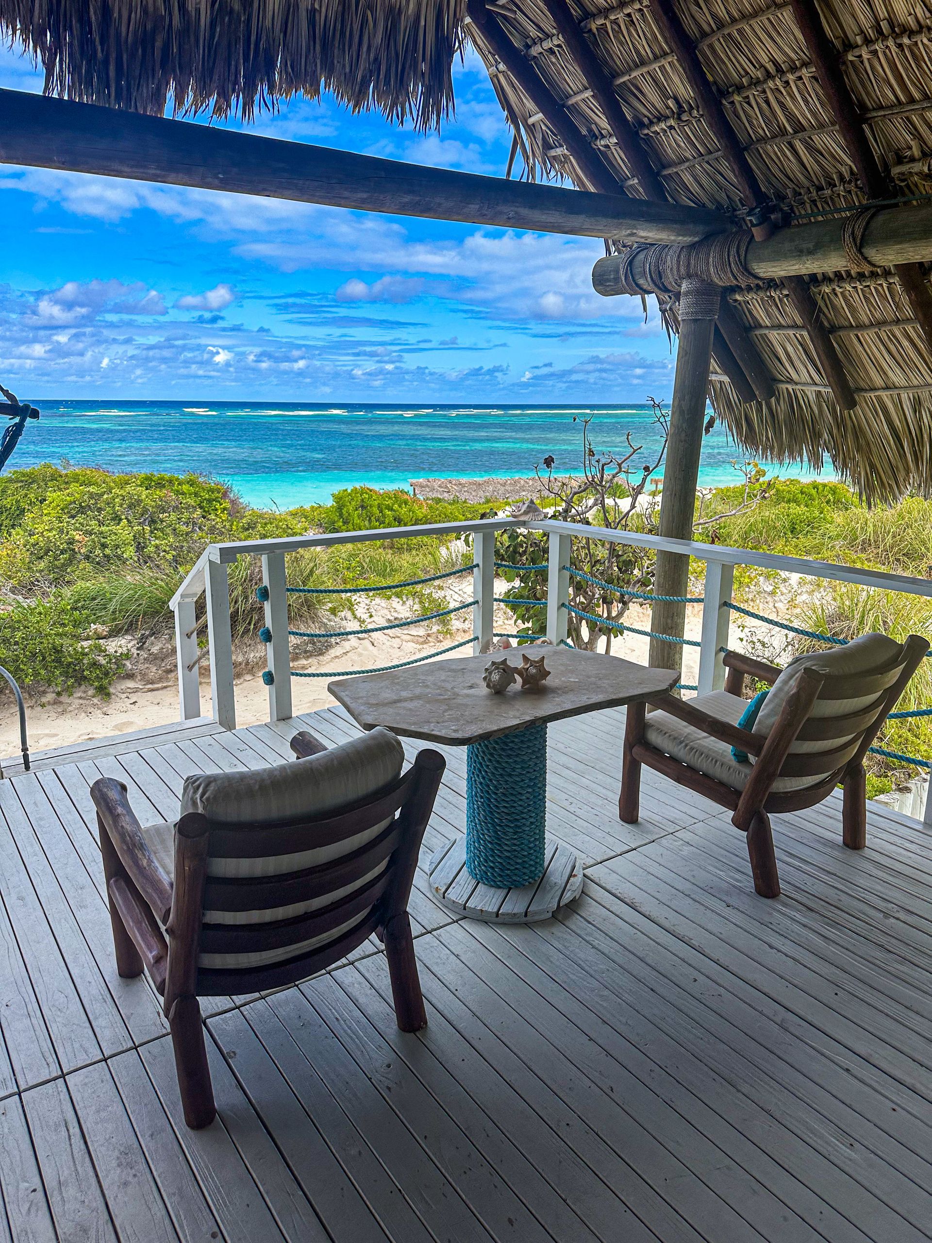 Beachside deck with chairs, table, ocean view. Turquoise water, blue sky, thatched roof.