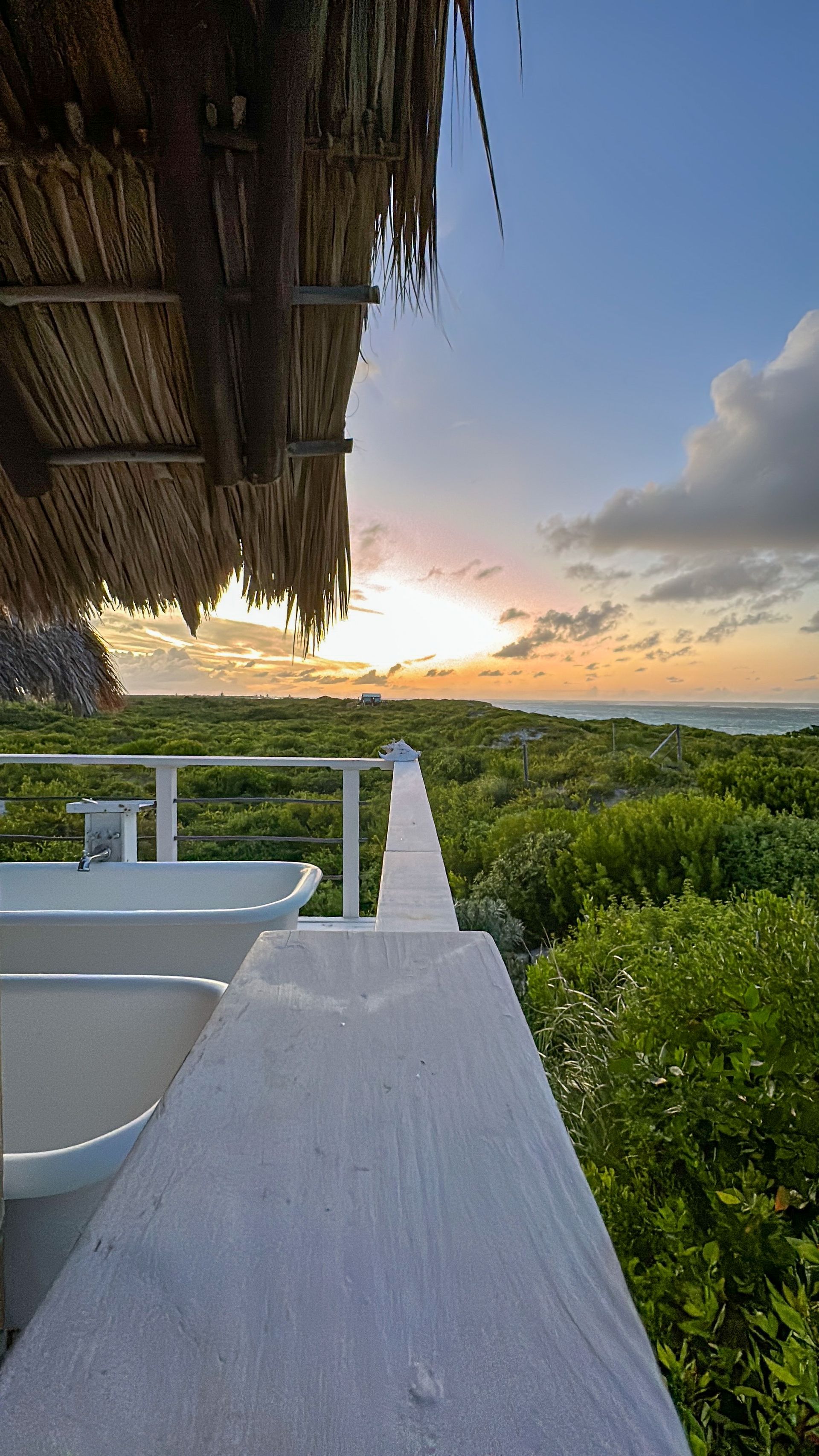 Sunrise over a lush landscape, viewed from a porch with white railing and thatched roof.