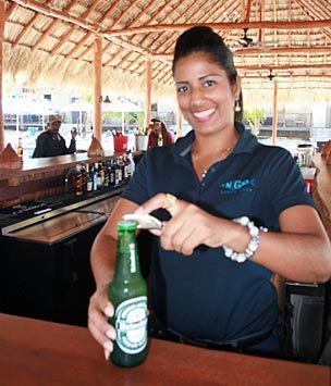 Woman in blue shirt opening a green beer bottle at a bar, smiling.
