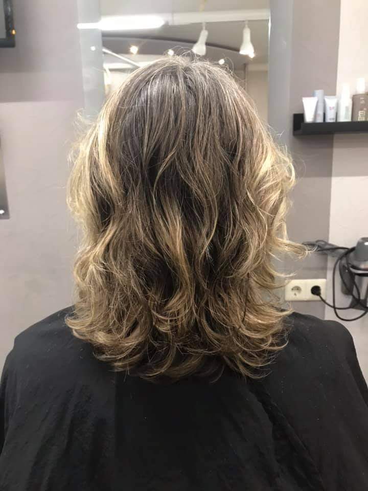 Eine Frau mit langem, welligem Haar sitzt in einem Friseursalon auf einem Stuhl vor einem Spiegel.