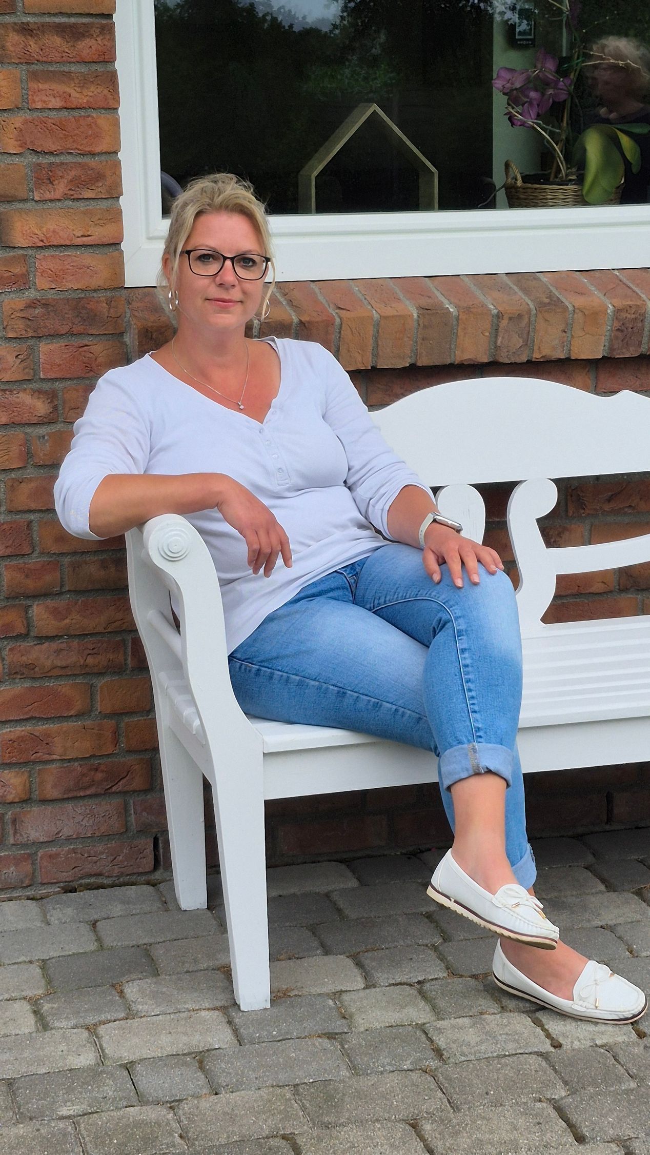 Eine Frau sitzt auf einer weißen Bank vor einem Backsteingebäude.