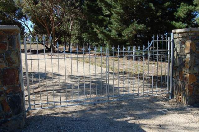 A metal gate is open to a dirt road with trees in the background.