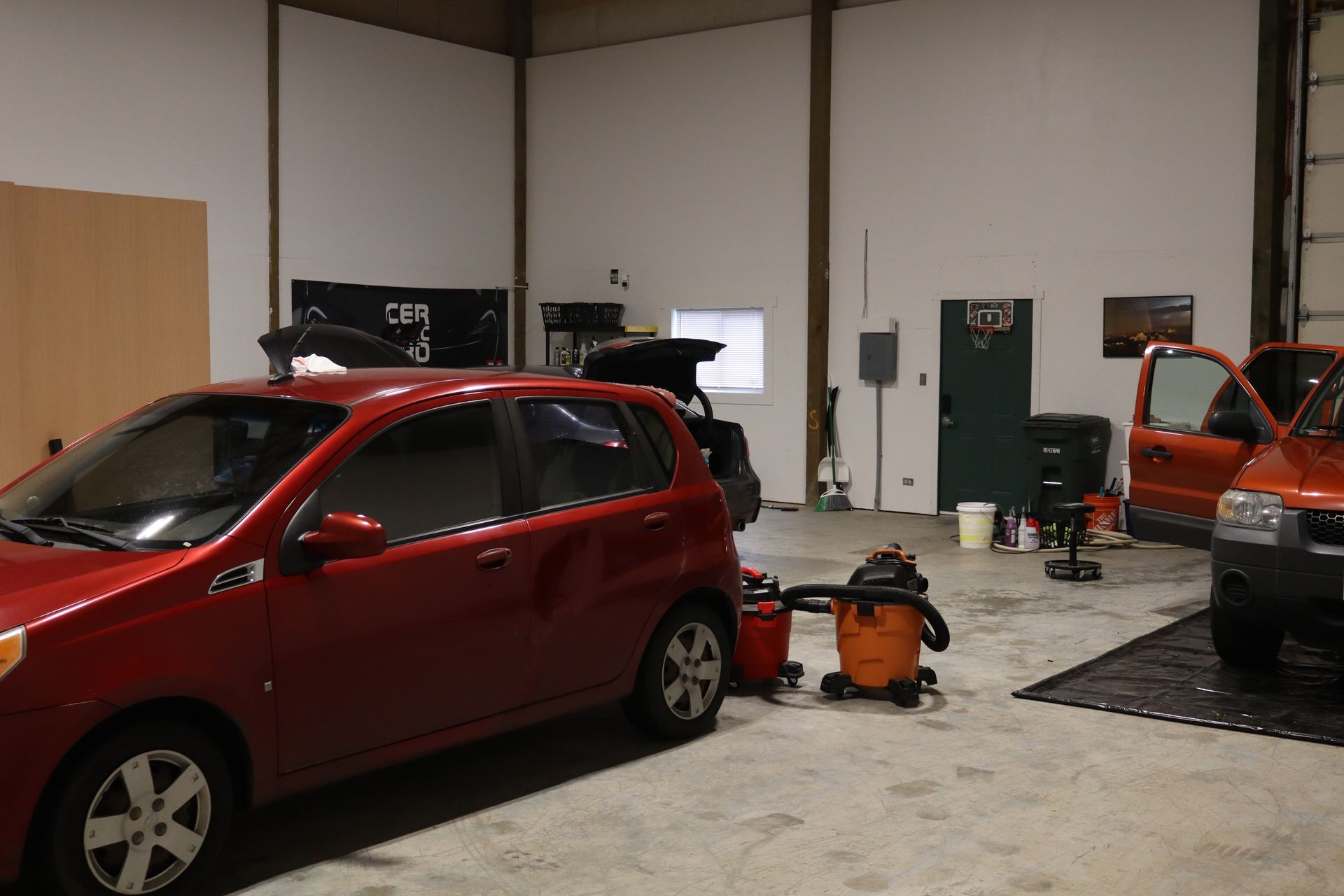 A red car is parked in a garage next to a vacuum cleaner.