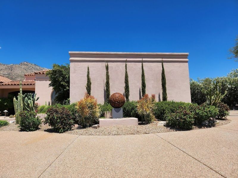 Tan stucco wall with cypress trees, bronze sphere sculpture, and desert landscaping under a blue sky.