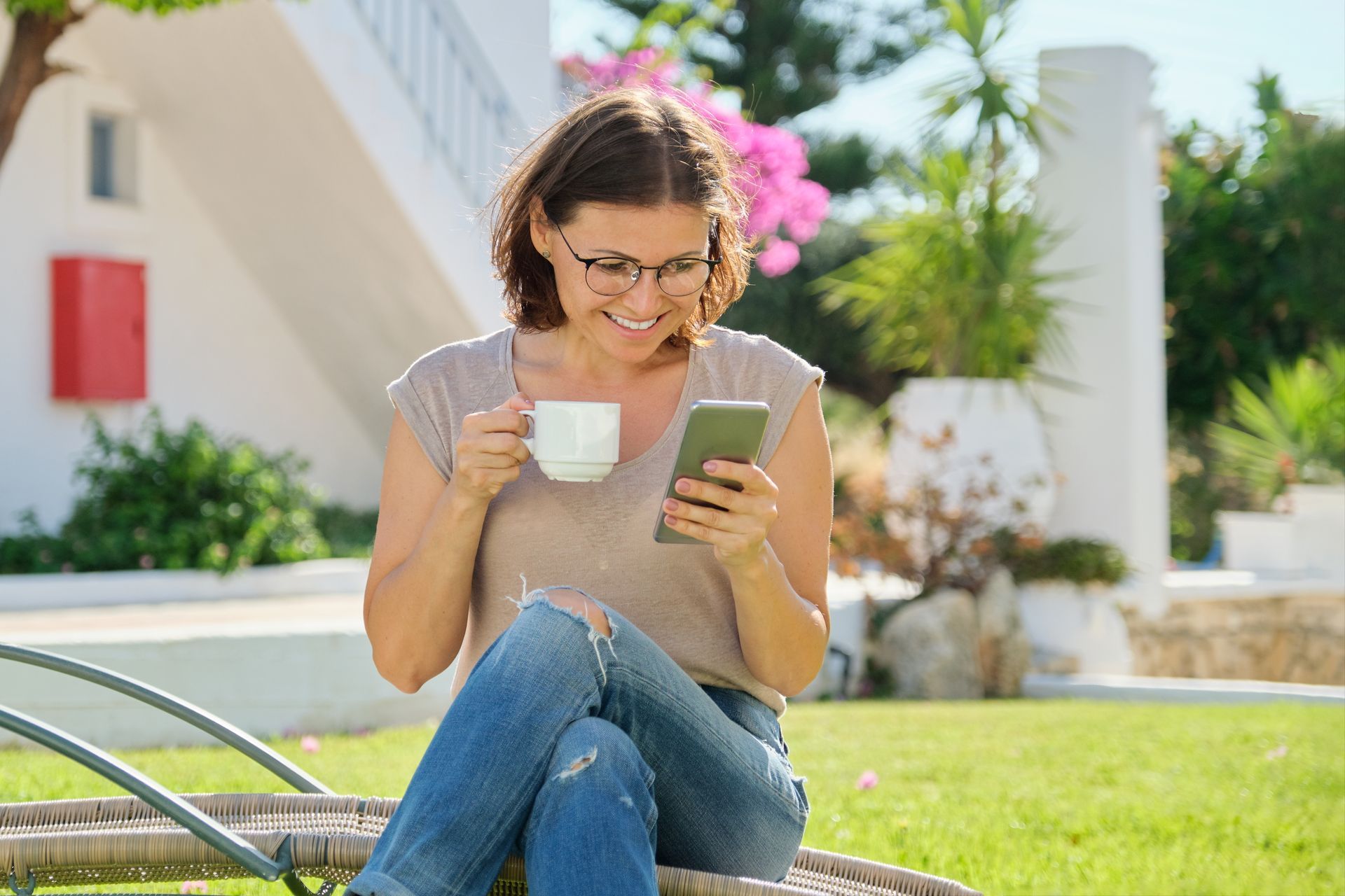 Woman with glasses, smiling, drinks coffee while looking at her phone outside on a sunny day.