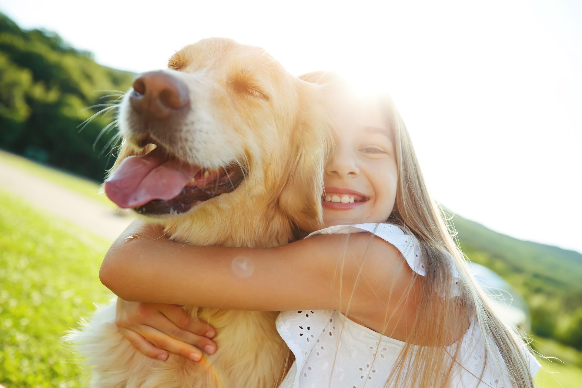 Girl hugging a golden retriever dog, smiling with sun in the background.