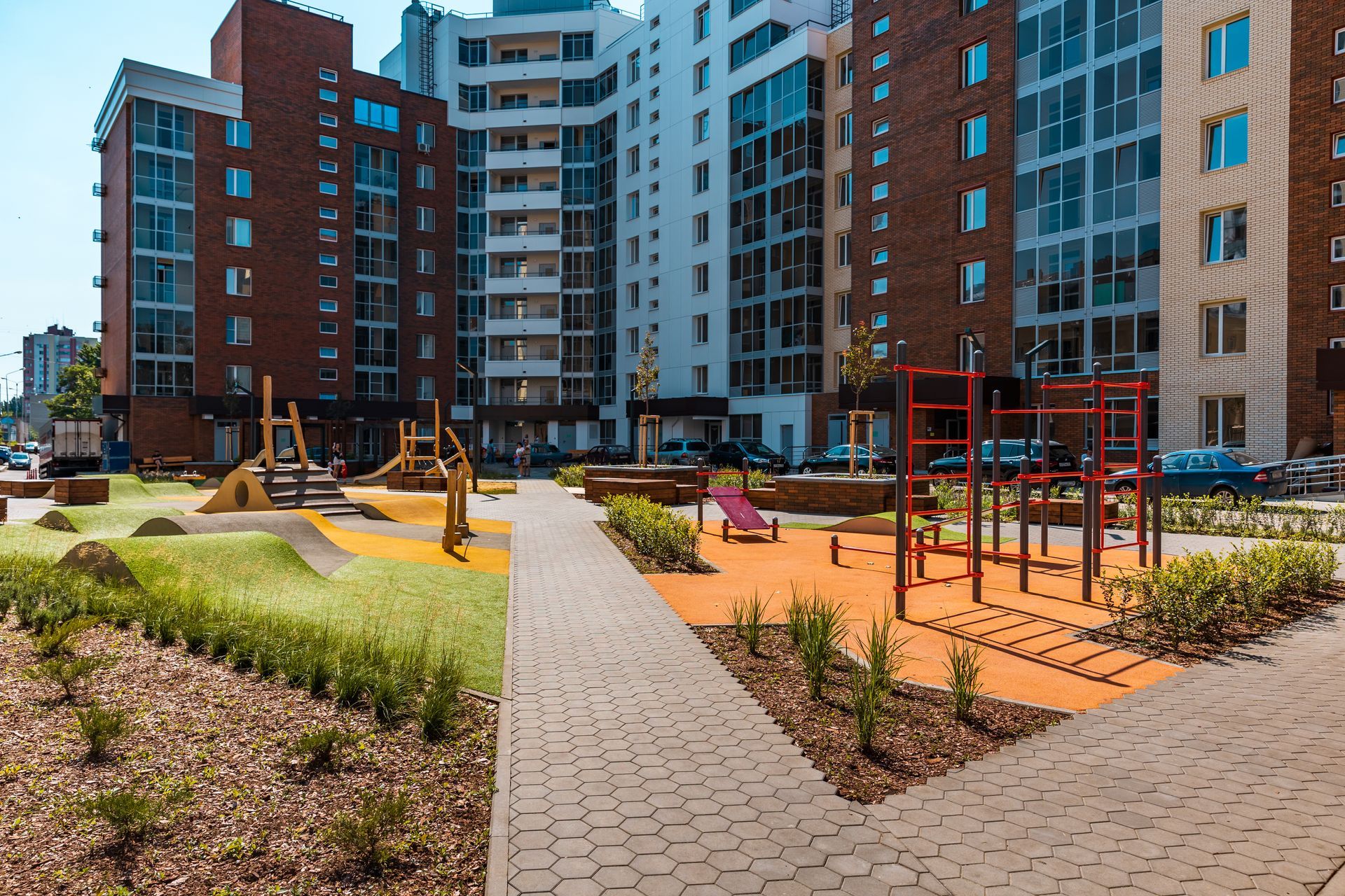 A courtyard playground with brick buildings. There is a paved path, colorful play equipment, and greenery.