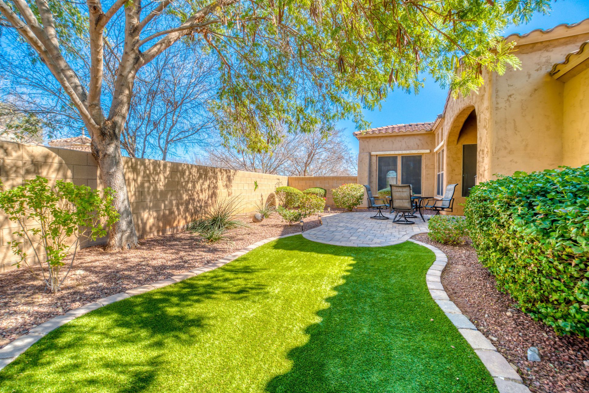 A backyard with green artificial turf, a patio with seating, and beige walls under a blue sky.